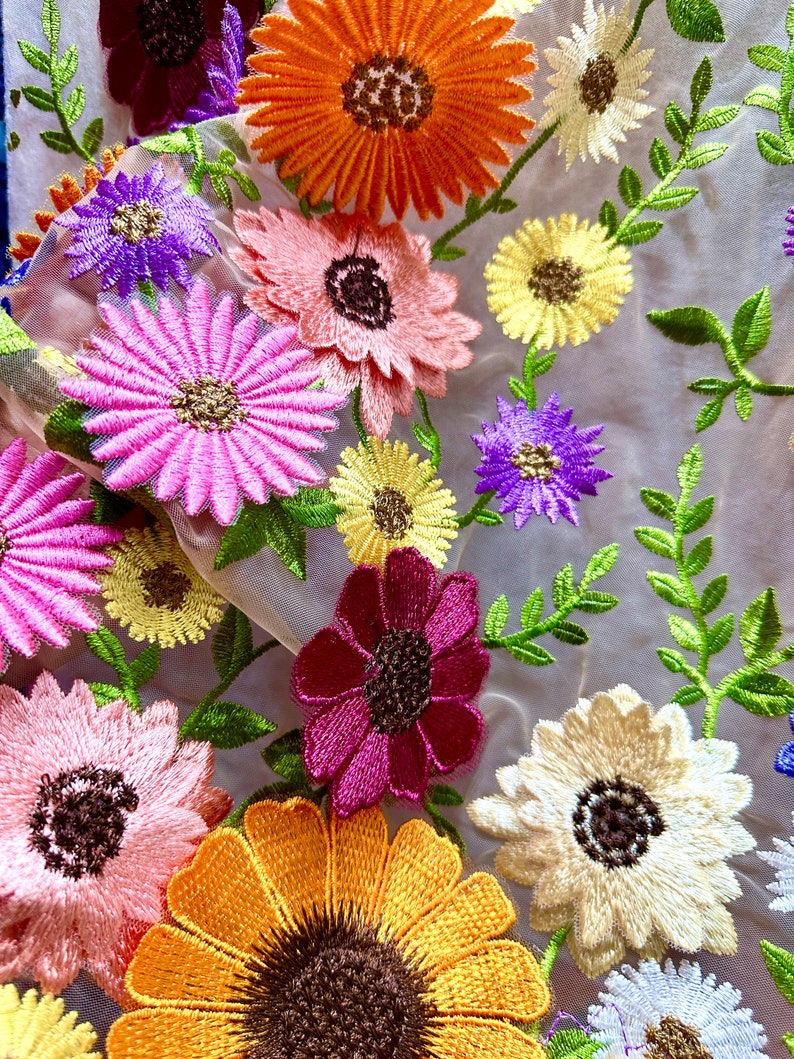 May include: A sheer fabric with embroidered flowers in various colors, including pink, purple, yellow, orange, and red. The flowers are arranged in a random pattern and are surrounded by green leaves.