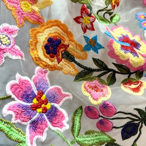 Puede incluir: Tela blanca transparente con flores bordadas de colores. Las flores son en tonos de rosa, morado, amarillo, naranja, rojo y verde. La tela tiene una textura delicada y aireada.