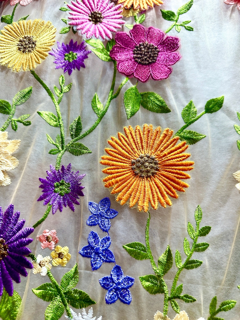 May include: A close-up of a beige mesh fabric with embroidered flowers in various colors, including yellow, orange, pink, purple, and blue. The flowers are arranged in a random pattern and are surrounded by green leaves.