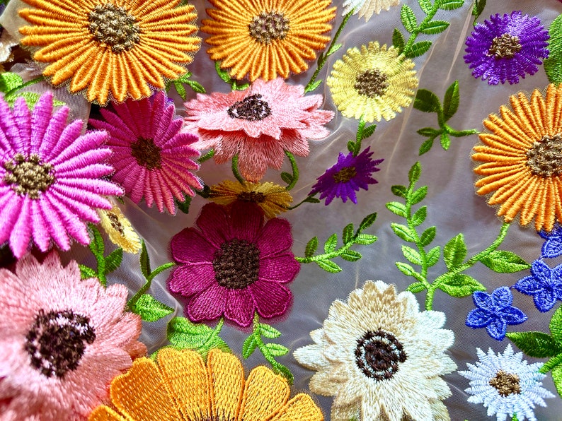 May include: A close-up of a sheer fabric with embroidered flowers in various colors, including pink, yellow, white, and purple. The flowers are arranged in a random pattern and have a delicate, feminine look.