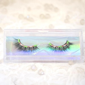 May include: A pair of false eyelashes in a clear plastic case. The lashes are black with green and pink accents. The case sits on a white lace surface with two white pearl-like beads.