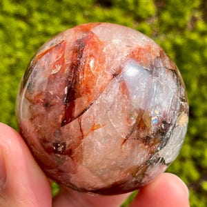 Może przedstawiać: Sfera z czerwonego i przezroczystego kryształu kwarcu o gładkiej, wypolerowanej powierzchni. Sfera jest trzymana w dłoni, a tło stanowi zielona powierzchnia mchu.