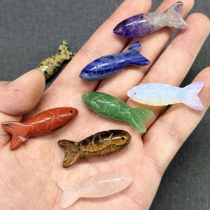 May include: A selection of small, carved stone fish in a variety of colours, including red, blue, green, and purple. The fish are held in a hand, showcasing their detailed craftsmanship and natural stone patterns. The collection includes a variety of stone types.