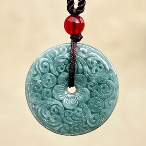 Peut inclure: Pendentif en jade vert clair orné de motifs floraux complexes. Il est suspendu à un cordon marron foncé avec une perle rouge. Le pendentif circulaire présente un trou central et est un bijou élégant.