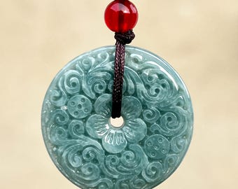May include: A jade pendant with intricate floral carvings. The pendant is a light teal color and is suspended from a dark brown cord with a red bead. The pendant has a circular shape with a hole in the center.