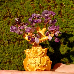 Könnte beinhalten: Ein dekorativer Amethyst-Edelsteinbaum mit goldfarbenem Sockel und Ästen. Der Baum ist mit kleinen, violetten Amethyststeinen und Kupferdraht verziert. Der Sockel ist ein goldfarbener Topf mit aufwendigen Details. Der Baum wird in einer Hand vor grünem Hintergrund gehalten.