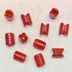 May include: A collection of red, carved beads in various shapes, including cylinders, bowties, and donut shapes. The beads have a textured surface and are displayed on a neutral background. These beads are suitable for jewelry making.