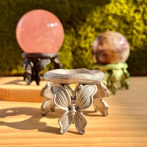 May include: A silver butterfly-shaped crystal ball holder is in the foreground. Behind it are two crystal balls on decorative stands. One is a pink sphere, and the other is a brown and purple sphere. The background is a blurred green.