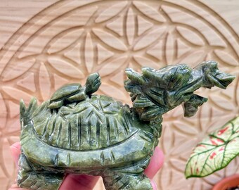 May include: A detailed green jade turtle figurine with a dragon-like head and a smaller turtle on its back. The sculpture has intricate carvings and a polished finish. The figurine is held in a hand, showcasing its size and detail.