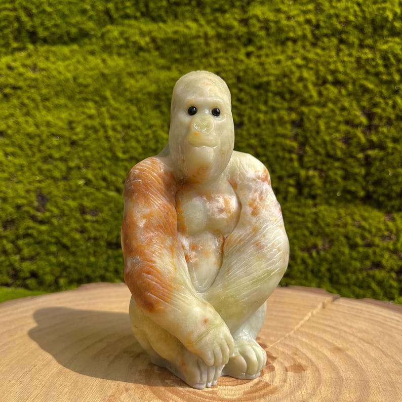 May include: A hand-carved stone sculpture of a gorilla sitting with its arms crossed. The sculpture is made of a light green stone with brown and orange markings. The gorilla has a realistic expression with large, dark eyes.