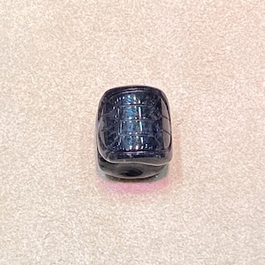 May include: A dark blue, barrel-shaped bead with a textured surface. The bead has a grid-like pattern and a glossy finish, reflecting light. The bead is approximately 0.75 inches in height and width.