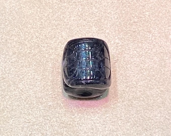 May include: A dark blue, barrel-shaped bead with a textured surface. The bead has a grid-like pattern and a glossy finish, reflecting light. The bead is approximately 0.75 inches in height and width.