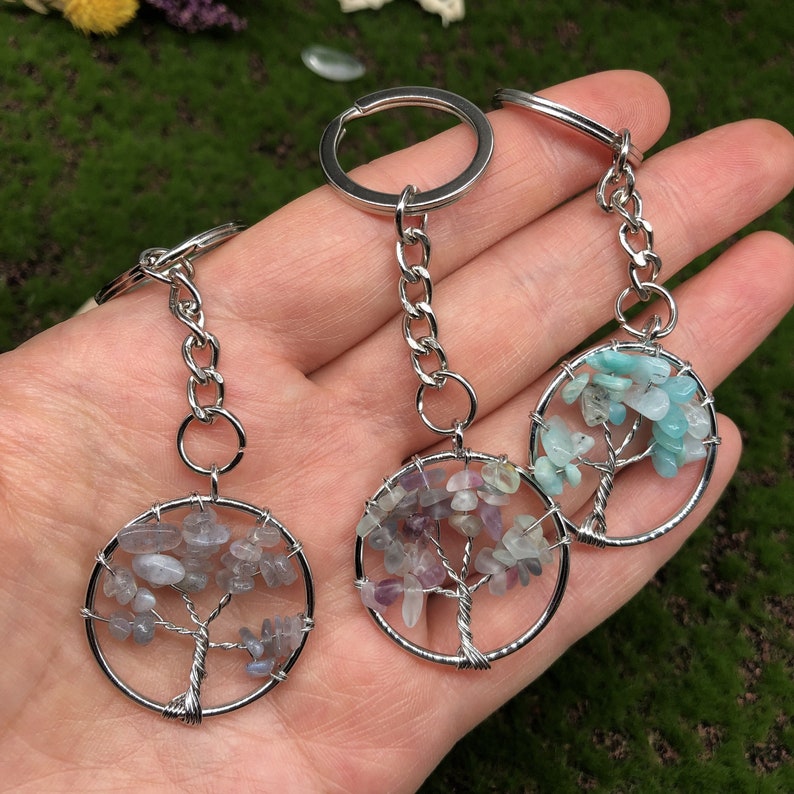 May include: Three silver tree of life keychains with different colored stones. The first keychain has light blue and green stones, the second has purple and pink stones, and the third has gray and blue stones.