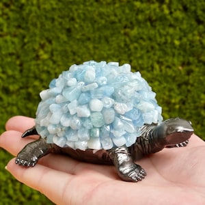 Puede incluir: Figura decorativa de tortuga con cuerpo y patas metálicas oscuras. El caparazón de la tortuga está hecho de piedras preciosas azul claro y blancas. La figura se sostiene en una mano sobre un fondo verde borroso.