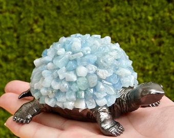May include: A decorative turtle figurine with a dark metallic body and legs. The turtle's shell is made of light blue and white gemstones. The figurine is held in a hand against a blurred green background.