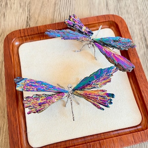 Pode incluir: Duas esculturas de libélulas com asas iridescentes em tons de roxo, azul e verde repousam sobre uma superfície creme dentro de uma bandeja de madeira. As libélulas têm corpos prateados e pernas delicadas.