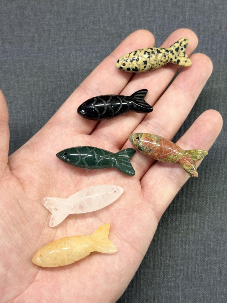 May include: Six carved stone fish figurines in a variety of colours and patterns. The fish are in shades of black, green, yellow, white, and speckled. The figurines are held in a person's hand against a grey background.