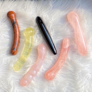May include: An assortment of crystal massage wands in various colors. The wands include a reddish-brown speckled wand, a yellow wand, a black wand, and several pink wands. The wands are smooth and curved.