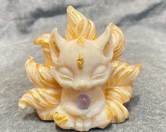 May include: A cream-colored fox figurine with nine golden-edged tails. The fox is in a seated position, holding a purple orb. Gold accents adorn the ears and forehead. The figurine is a decorative item.