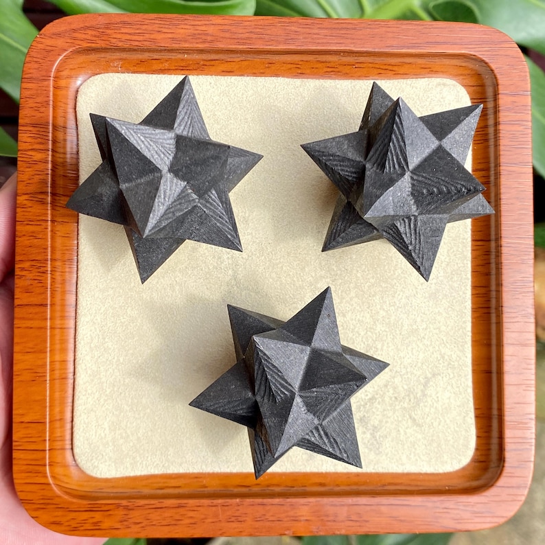 May include: Three dark gray, star-shaped objects with geometric facets are displayed on a beige surface within a wooden tray. The stars have a textured appearance, and the tray has a polished, warm-toned wood finish.