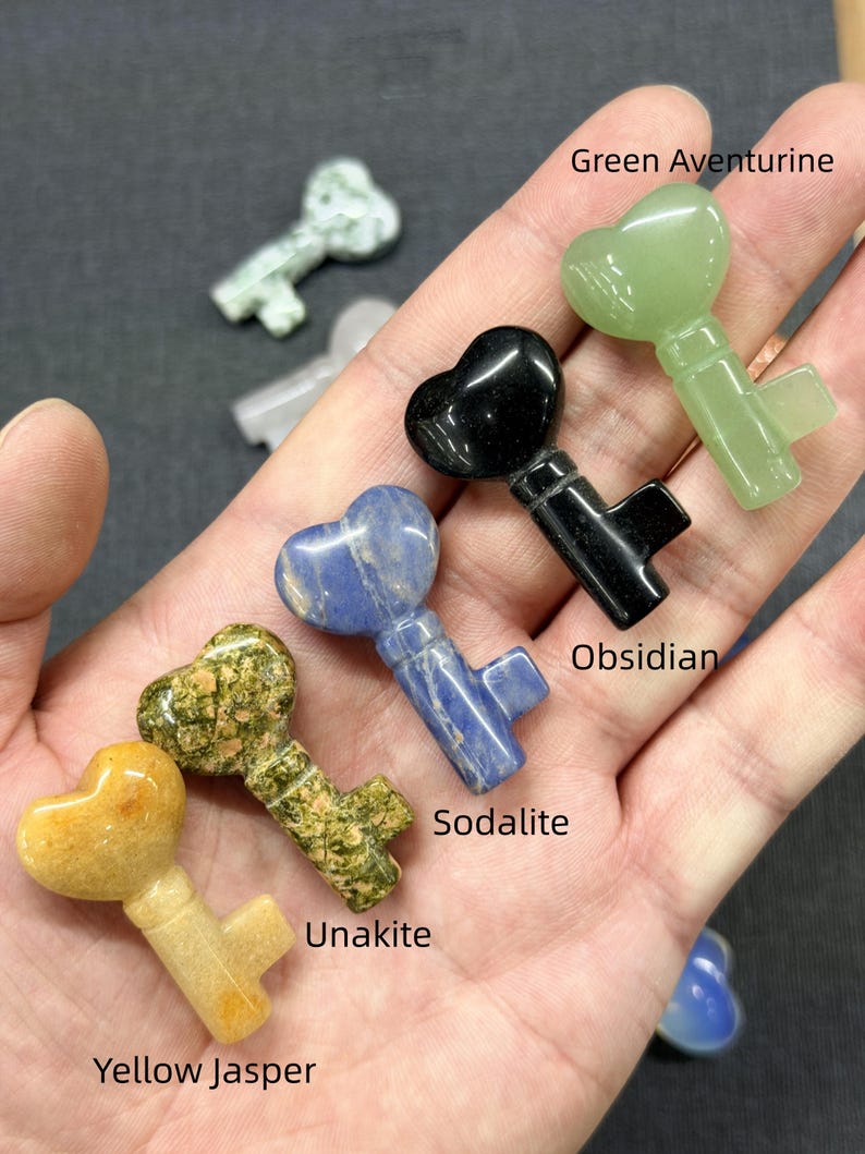 Op de afbeelding: Een verzameling edelstenen sleutelhangers, waaronder gele jaspis, unakiet, sodaliet, obsidiaan en groene aventurijn. Elke sleutel heeft een hartvormige bovenkant en een sleutelvormige onderkant. De edelstenen vari&euml;ren in kleur.