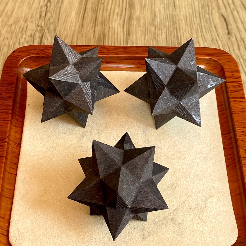 May include: Three dark gray, geometric star-shaped objects are displayed on a wooden tray with a light beige surface. The stars have a faceted design, creating a textured appearance. The tray has a dark brown border.