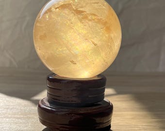 May include: A large, clear, yellow citrine crystal sphere on a dark brown wooden stand. The sphere is polished and has a slight iridescent sheen.
