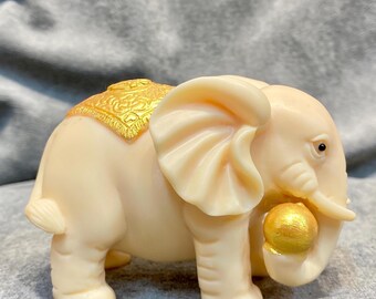 May include: Ivory-colored elephant figurine with gold accents. The elephant holds a gold ball in its trunk and has a decorative gold saddle. The figurine is a detailed, decorative item.