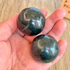 Può includere: Due sfere lucide verde scuro con una superficie riflettente sono tenute in una mano. Le sfere hanno una forma liscia e arrotondata e sembrano essere fatte di una pietra naturale o minerale. Il colore è un verde intenso e ricco.