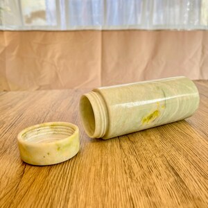 May include: A cylindrical container with a matching lid, crafted from a light green and yellow veined stone. The container is open, revealing the interior. The lid sits beside the container on a wooden surface.
