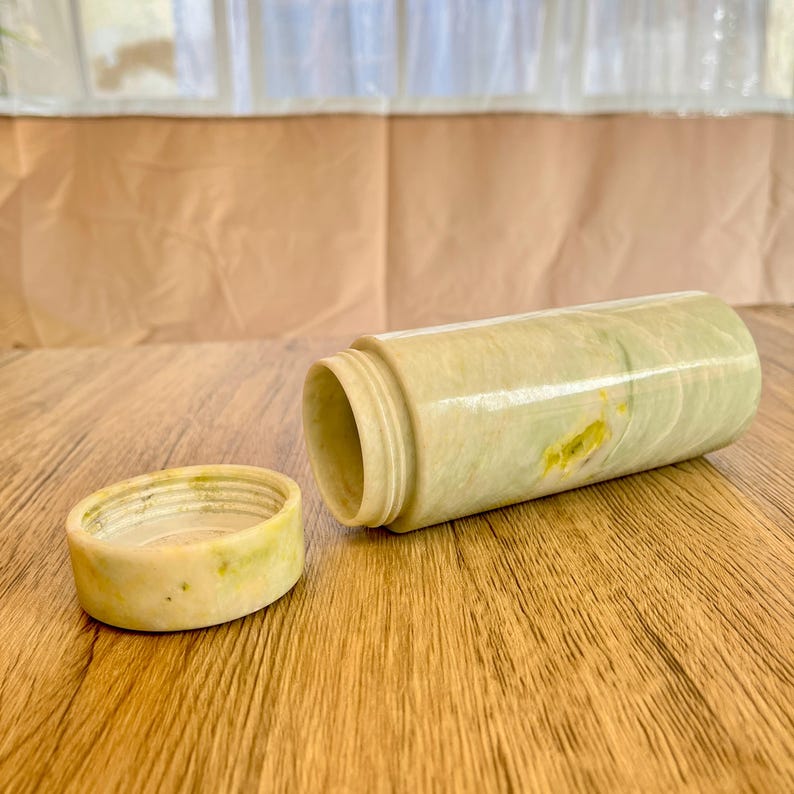 May include: A cylindrical container with a matching lid, crafted from a light green and yellow veined stone. The container is open, revealing the interior. The lid sits beside the container on a wooden surface.