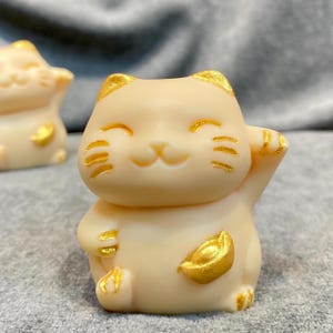 May include: A cream-colored Maneki Neko figurine with gold accents. The lucky cat is in a welcoming pose, with one paw raised. Gold details include the ears, coin, and facial markings. The figurine is a symbol of good fortune and prosperity.