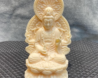May include: Ivory-colored statue of a seated figure with a serene expression, adorned with intricate details. The figure is seated on a lotus flower base, with a halo-like design behind the head. The statue is likely a representation of a religious or spiritual figure.