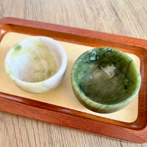 May include: Two small, round, stone bowls in shades of green and white sit on a wooden tray. The bowls have a smooth, polished finish and a slight indentation in the center. The tray has a dark brown border and a light tan interior.