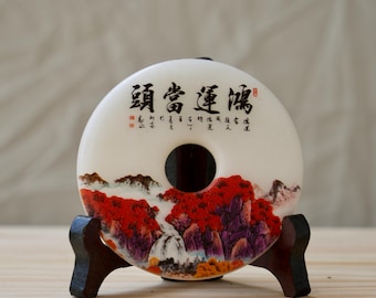 May include: A white ceramic donut-shaped ornament with a red and purple landscape design and Chinese calligraphy. The ornament is on a wooden stand.