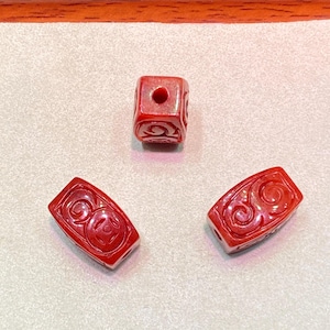 May include: Three red carved beads with a swirling design. One bead is cube-shaped with a hole, and the other two are rectangular. The beads are displayed on a light gray surface.