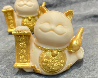 May include: Two cream-colored Maneki Neko figurines with gold accents. The cats are smiling and holding a gold scroll and a gold bottle. The figurines have gold collars and are on a gray surface.
