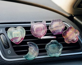 May include: Six colorful car air freshener charms in various shapes, including a heart, a cat, a flower, a crescent moon, a hexagon, and a flower. The charms are made of iridescent glass and are hanging from the air vent of a car.