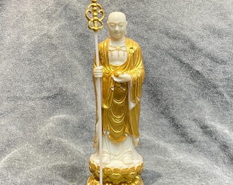 May include: A white and gold statue of a Buddhist monk holding a staff. The monk is wearing a gold robe and is standing on a gold lotus flower base. The staff has a decorative gold top.