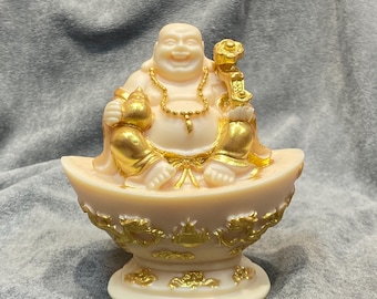 May include: A cream-colored Buddha statue with gold accents. The smiling figure is seated on a gold-detailed base, holding a gold object in one hand and a gold sack in the other. A gold necklace adorns the Buddha's neck.