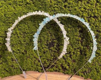 Op de afbeelding: Twee zilveren haarbanden versierd met lichtblauwe en witte edelsteenaccenten. De haarbanden worden getoond tegen een groene mosachtige achtergrond.