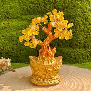 May include: A golden-colored decorative tree with yellow crystal leaves. The tree sits in a gold-colored pot with Chinese characters and coin details. The tree is on a wooden surface with a green moss background.