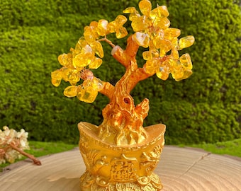 May include: A golden-colored decorative tree with yellow crystal leaves. The tree sits in a gold-colored pot with Chinese characters and coin details. The tree is on a wooden surface with a green moss background.