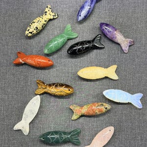 May include: A selection of carved stone fish ornaments in diverse colours and patterns. The fish feature orange, green, yellow, black, and speckled designs. Each fish has a detailed fin and tail.