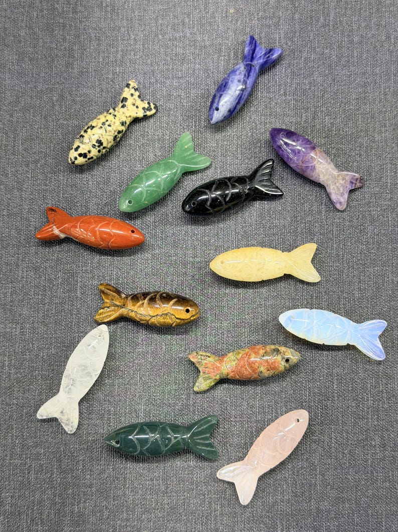 May include: A selection of carved stone fish ornaments in diverse colours and patterns. The fish feature orange, green, yellow, black, and speckled designs. Each fish has a detailed fin and tail.