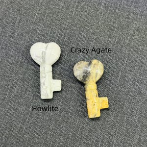 Op de afbeelding: Twee hartvormige sleutelhangers. E&eacute;n is wit met grijze adering, met het label "Howlite". De andere is geel, bruin en grijs, met het label "Crazy Agate". Beide liggen op een grijze, getextureerde ondergrond.
