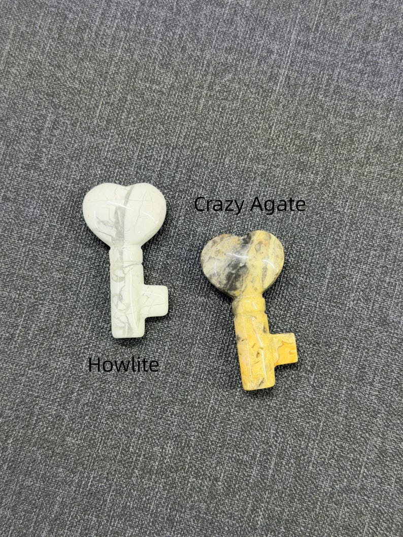 Op de afbeelding: Twee hartvormige sleutelhangers. E&eacute;n is wit met grijze adering, met het label "Howlite". De andere is geel, bruin en grijs, met het label "Crazy Agate". Beide liggen op een grijze, getextureerde ondergrond.