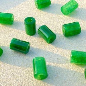 Könnte beinhalten: Eine Ansammlung leuchtend grüner, zylindrischer Perlen mit polierter, glänzender Oberfläche. Jede Perle hat ein Loch in der Mitte und ist auf einer hellen Oberfläche verstreut. Die Perlen sind etwa 1,3 cm lang.