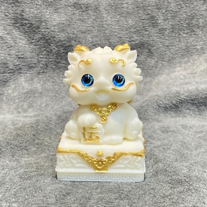 May include: A white figurine of a mythical creature with blue eyes and gold accents. The figurine is seated on a decorative base with gold trim. The creature holds a gold object. The overall design is detailed and ornate.