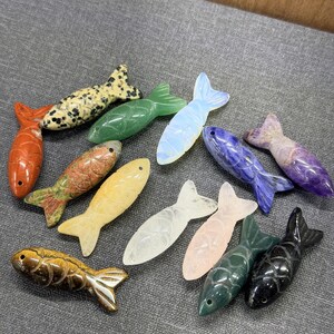 May include: A selection of carved stone fish in diverse colours and patterns. The fish include orange, green, yellow, white, pink, blue, and black. Some have speckled patterns, while others are solid colours. The fish are arranged on a grey surface.